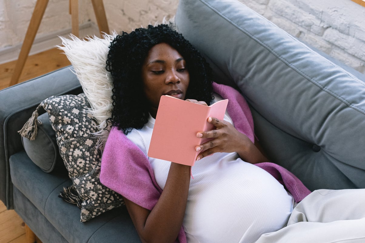 Black pregnant woman writing in notebook on couch